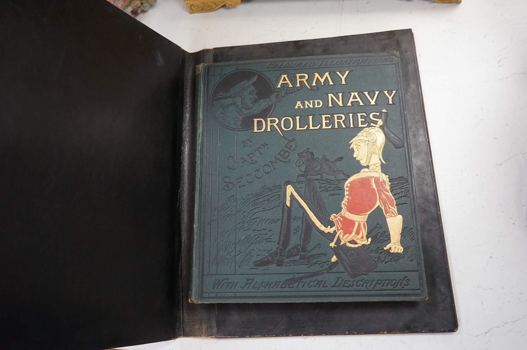 An olive and marquetry desk blotter, Duke of Wellington on horseback and his coat of arms, together with a cloth and gilt book, Army and Navy Drolleries by Captain Seccombe